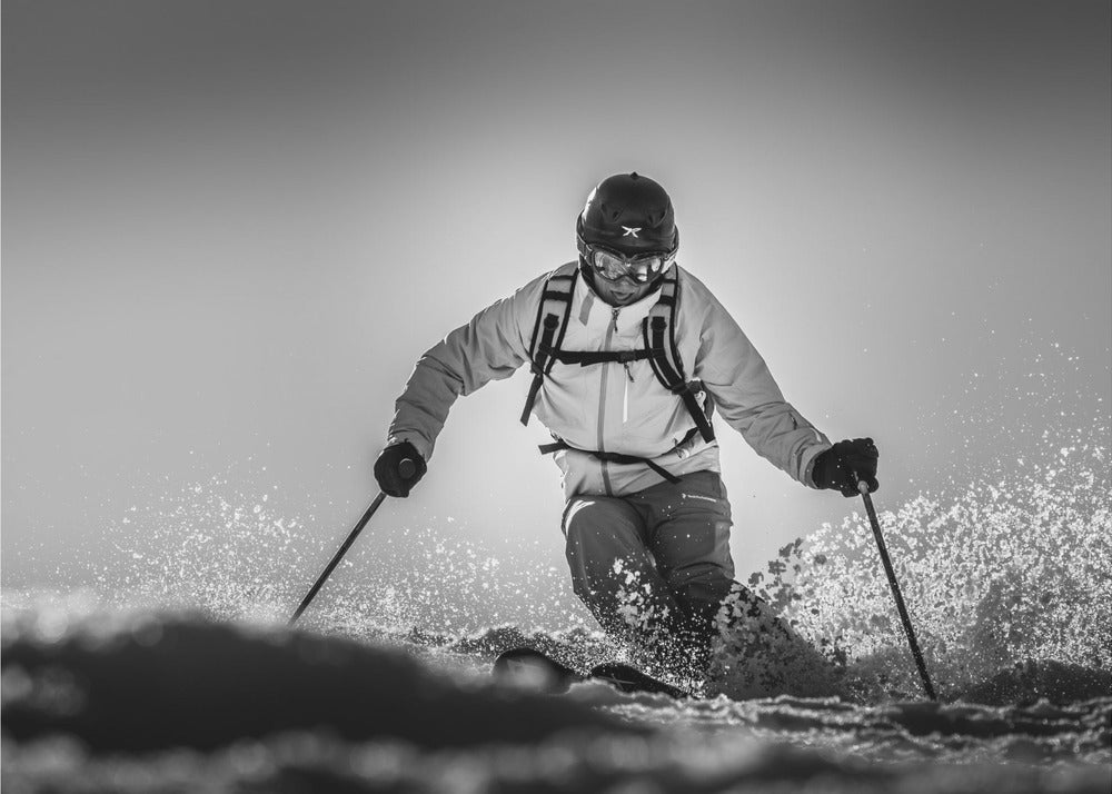 A black and white action photograph of a skier in full gear, leaning into a turn and spraying powder snow. The skier is backlit by the sun, creating a high-contrast, dramatic effect. Print