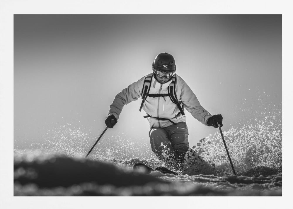 A black and white action photograph of a skier in full gear, leaning into a turn and spraying powder snow. The skier is backlit by the sun, creating a high-contrast, dramatic effect. Print
