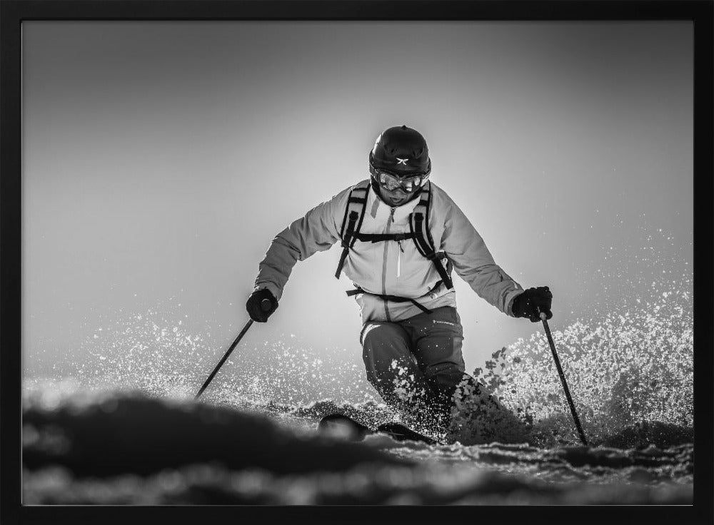 A black and white action photograph of a skier in full gear, leaning into a turn and spraying powder snow. The skier is backlit by the sun, creating a high-contrast, dramatic effect. Print