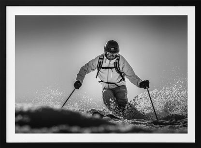 A black and white action photograph of a skier in full gear, leaning into a turn and spraying powder snow. The skier is backlit by the sun, creating a high-contrast, dramatic effect. Print
