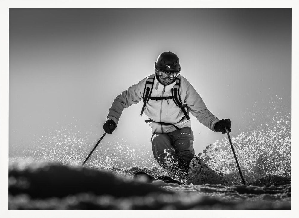 A black and white action photograph of a skier in full gear, leaning into a turn and spraying powder snow. The skier is backlit by the sun, creating a high-contrast, dramatic effect. Print