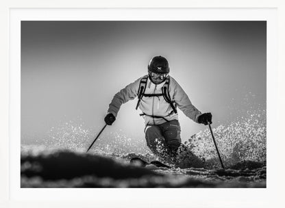 A black and white action photograph of a skier in full gear, leaning into a turn and spraying powder snow. The skier is backlit by the sun, creating a high-contrast, dramatic effect. Print