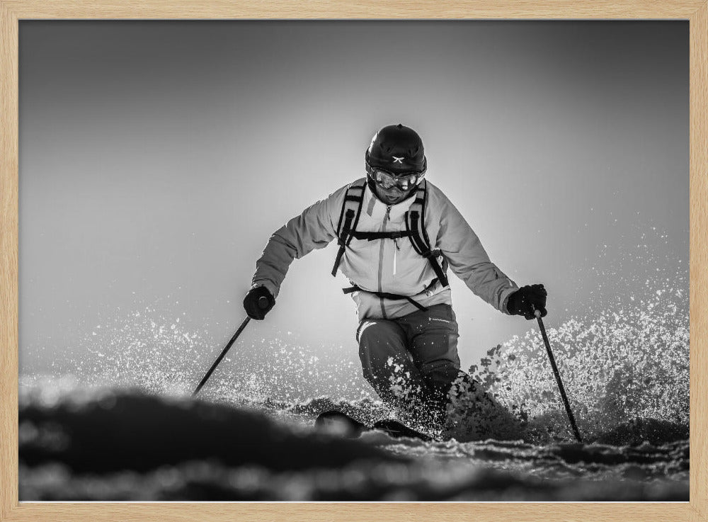 A black and white action photograph of a skier in full gear, leaning into a turn and spraying powder snow. The skier is backlit by the sun, creating a high-contrast, dramatic effect. Print