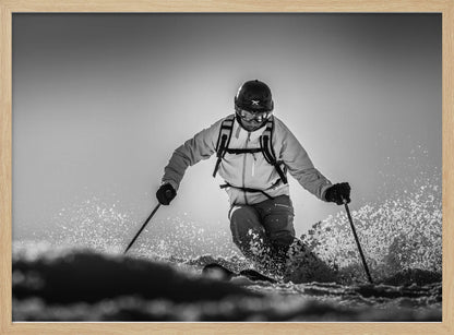 A black and white action photograph of a skier in full gear, leaning into a turn and spraying powder snow. The skier is backlit by the sun, creating a high-contrast, dramatic effect. Print