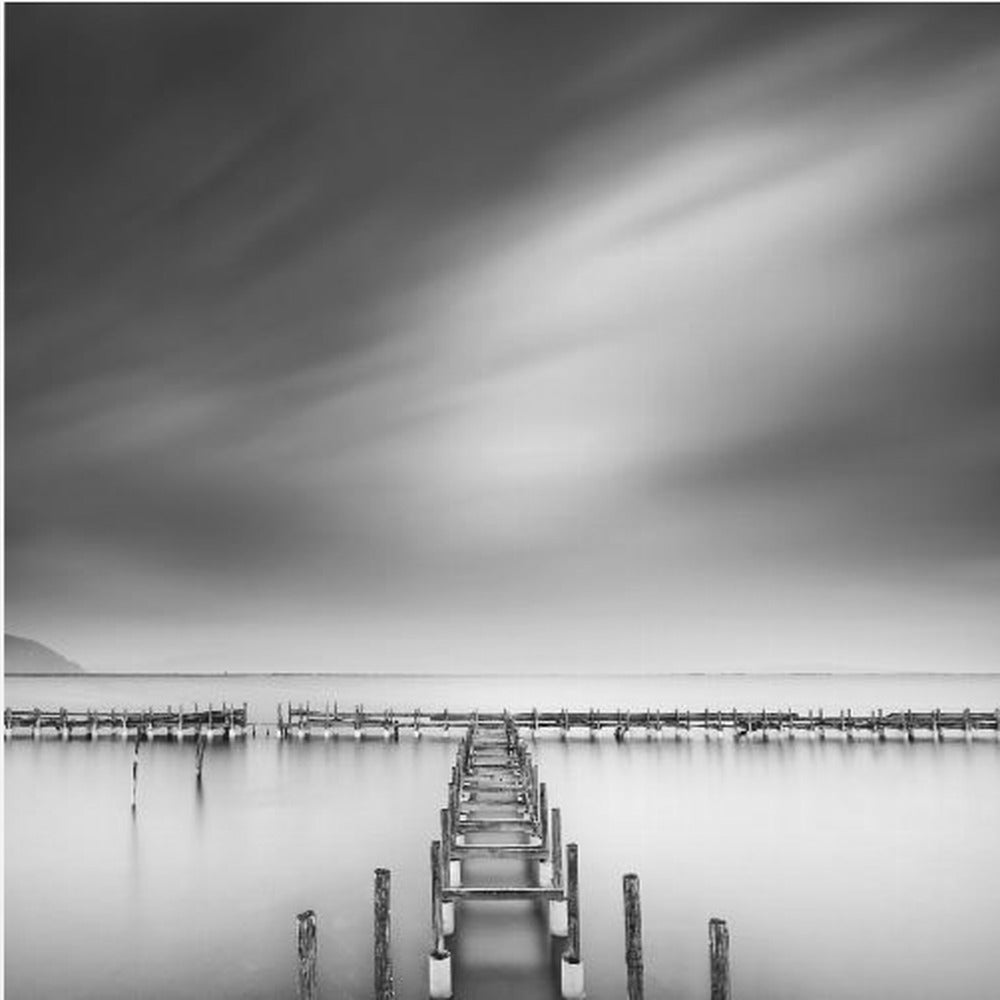 A framed, square black and white long-exposure photograph of a dilapidated wooden pier extending into calm, misty water. The sky is filled with soft, streaked clouds, creating a serene and melancholic mood. Wall Art