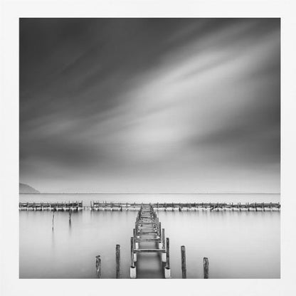 A framed, square black and white long-exposure photograph of a dilapidated wooden pier extending into calm, misty water. The sky is filled with soft, streaked clouds, creating a serene and melancholic mood. Wall Art