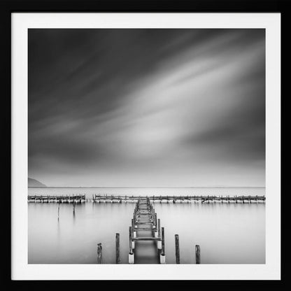 A framed, square black and white long-exposure photograph of a dilapidated wooden pier extending into calm, misty water. The sky is filled with soft, streaked clouds, creating a serene and melancholic mood. Wall Art
