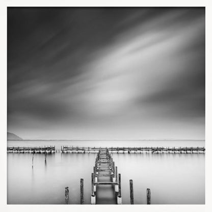 A framed, square black and white long-exposure photograph of a dilapidated wooden pier extending into calm, misty water. The sky is filled with soft, streaked clouds, creating a serene and melancholic mood. Wall Art