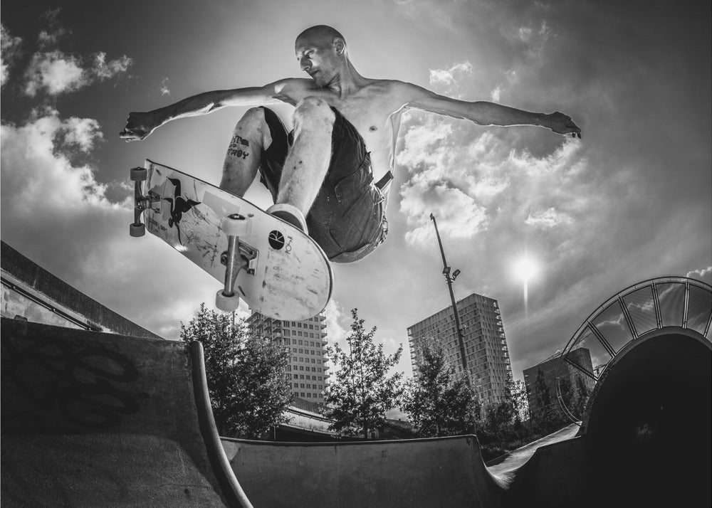 A dramatic low-angle, black and white photograph of a shirtless skateboarder caught in mid-air above a skatepark ramp. His arms are spread wide for balance as the sun flares brightly behind him, set against a cloudy urban sky with high-rise buildings in the distance. The image is enclosed in a silver frame. Wall Art