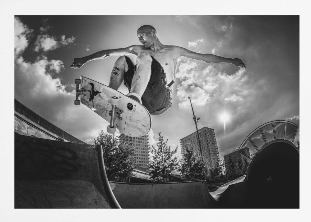 A dramatic low-angle, black and white photograph of a shirtless skateboarder caught in mid-air above a skatepark ramp. His arms are spread wide for balance as the sun flares brightly behind him, set against a cloudy urban sky with high-rise buildings in the distance. The image is enclosed in a silver frame. Wall Art