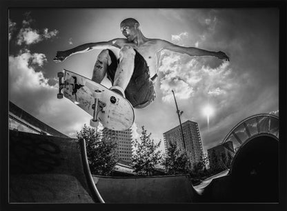 A dramatic low-angle, black and white photograph of a shirtless skateboarder caught in mid-air above a skatepark ramp. His arms are spread wide for balance as the sun flares brightly behind him, set against a cloudy urban sky with high-rise buildings in the distance. The image is enclosed in a silver frame. Wall Art