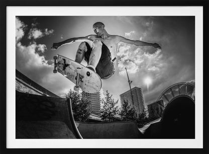 A dramatic low-angle, black and white photograph of a shirtless skateboarder caught in mid-air above a skatepark ramp. His arms are spread wide for balance as the sun flares brightly behind him, set against a cloudy urban sky with high-rise buildings in the distance. The image is enclosed in a silver frame. Wall Art