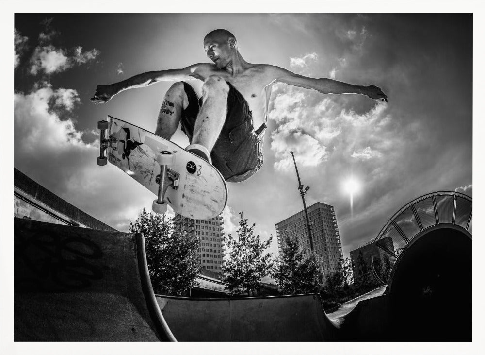A dramatic low-angle, black and white photograph of a shirtless skateboarder caught in mid-air above a skatepark ramp. His arms are spread wide for balance as the sun flares brightly behind him, set against a cloudy urban sky with high-rise buildings in the distance. The image is enclosed in a silver frame. Wall Art