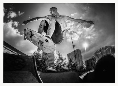 A dramatic low-angle, black and white photograph of a shirtless skateboarder caught in mid-air above a skatepark ramp. His arms are spread wide for balance as the sun flares brightly behind him, set against a cloudy urban sky with high-rise buildings in the distance. The image is enclosed in a silver frame. Wall Art