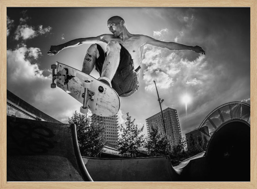 A dramatic low-angle, black and white photograph of a shirtless skateboarder caught in mid-air above a skatepark ramp. His arms are spread wide for balance as the sun flares brightly behind him, set against a cloudy urban sky with high-rise buildings in the distance. The image is enclosed in a silver frame. Wall Art