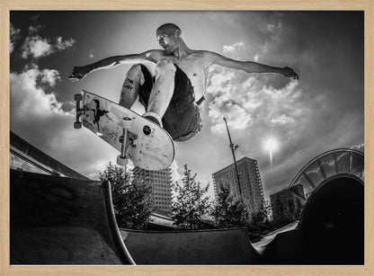 A dramatic low-angle, black and white photograph of a shirtless skateboarder caught in mid-air above a skatepark ramp. His arms are spread wide for balance as the sun flares brightly behind him, set against a cloudy urban sky with high-rise buildings in the distance. The image is enclosed in a silver frame. Wall Art