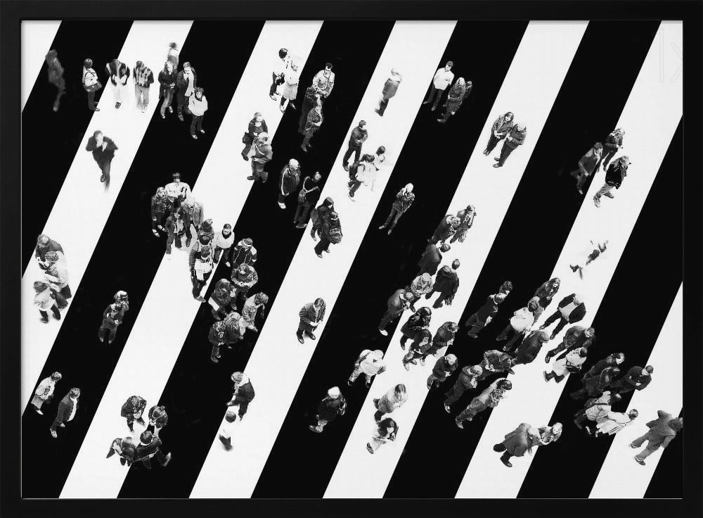 A striking black and white aerial photograph of dozens of people walking across a large, diagonally striped crosswalk. The high-contrast, graphic pattern of black and white stripes dominates the scene, with the small figures of pedestrians scattered across it, creating a sense of movement and urban anonymity. The image is presented within a simple silver frame. Print