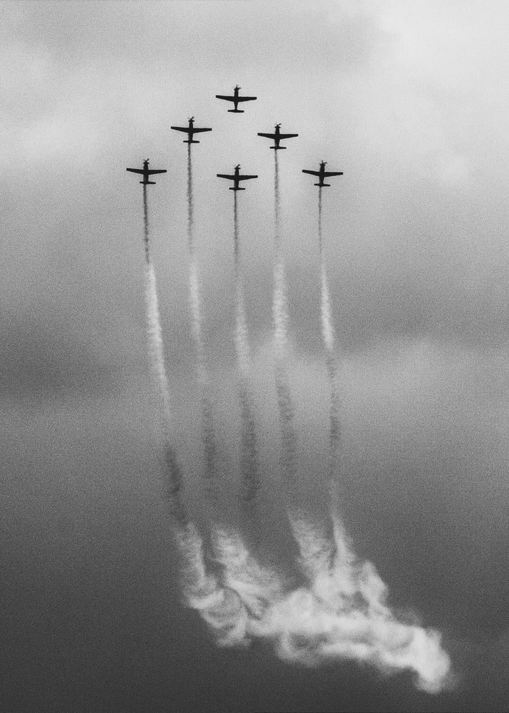 A grainy black and white photo of six airplanes flying in a V-formation against a cloudy sky. Each plane leaves a long, white smoke trail that billows at the bottom of the frame. The image is presented within a black border. Print