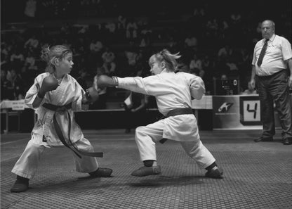 A black and white photograph of two young girls in a karate competition. They are wearing white gis, black belts, and boxing gloves. One girl is in the middle of throwing a punch at her opponent. In the background, a referee stands watching the match, and spectators are blurred in the stands. The image is presented within a silver-colored frame. Print