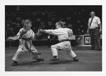 A black and white photograph of two young girls in a karate competition. They are wearing white gis, black belts, and boxing gloves. One girl is in the middle of throwing a punch at her opponent. In the background, a referee stands watching the match, and spectators are blurred in the stands. The image is presented within a silver-colored frame. Print