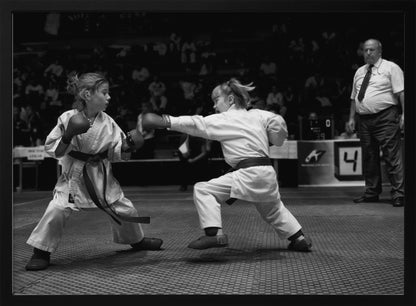 A black and white photograph of two young girls in a karate competition. They are wearing white gis, black belts, and boxing gloves. One girl is in the middle of throwing a punch at her opponent. In the background, a referee stands watching the match, and spectators are blurred in the stands. The image is presented within a silver-colored frame. Print