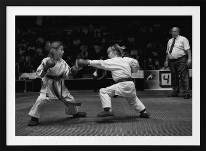 A black and white photograph of two young girls in a karate competition. They are wearing white gis, black belts, and boxing gloves. One girl is in the middle of throwing a punch at her opponent. In the background, a referee stands watching the match, and spectators are blurred in the stands. The image is presented within a silver-colored frame. Print