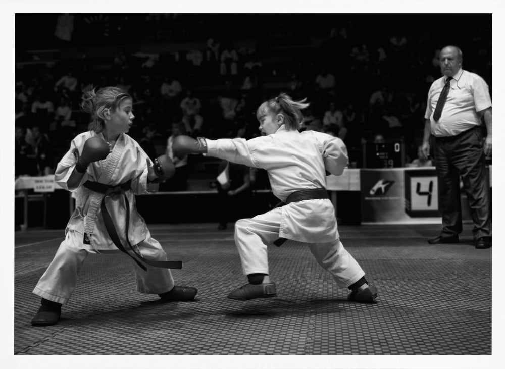 A black and white photograph of two young girls in a karate competition. They are wearing white gis, black belts, and boxing gloves. One girl is in the middle of throwing a punch at her opponent. In the background, a referee stands watching the match, and spectators are blurred in the stands. The image is presented within a silver-colored frame. Print