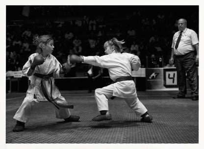 A black and white photograph of two young girls in a karate competition. They are wearing white gis, black belts, and boxing gloves. One girl is in the middle of throwing a punch at her opponent. In the background, a referee stands watching the match, and spectators are blurred in the stands. The image is presented within a silver-colored frame. Print