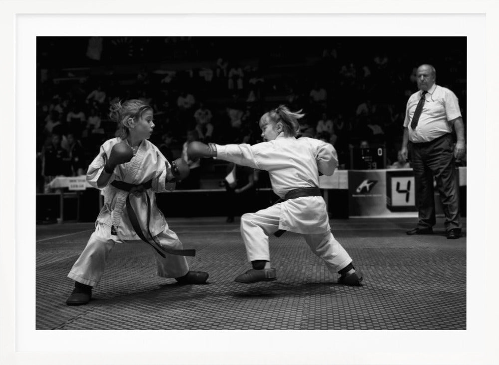 A black and white photograph of two young girls in a karate competition. They are wearing white gis, black belts, and boxing gloves. One girl is in the middle of throwing a punch at her opponent. In the background, a referee stands watching the match, and spectators are blurred in the stands. The image is presented within a silver-colored frame. Print