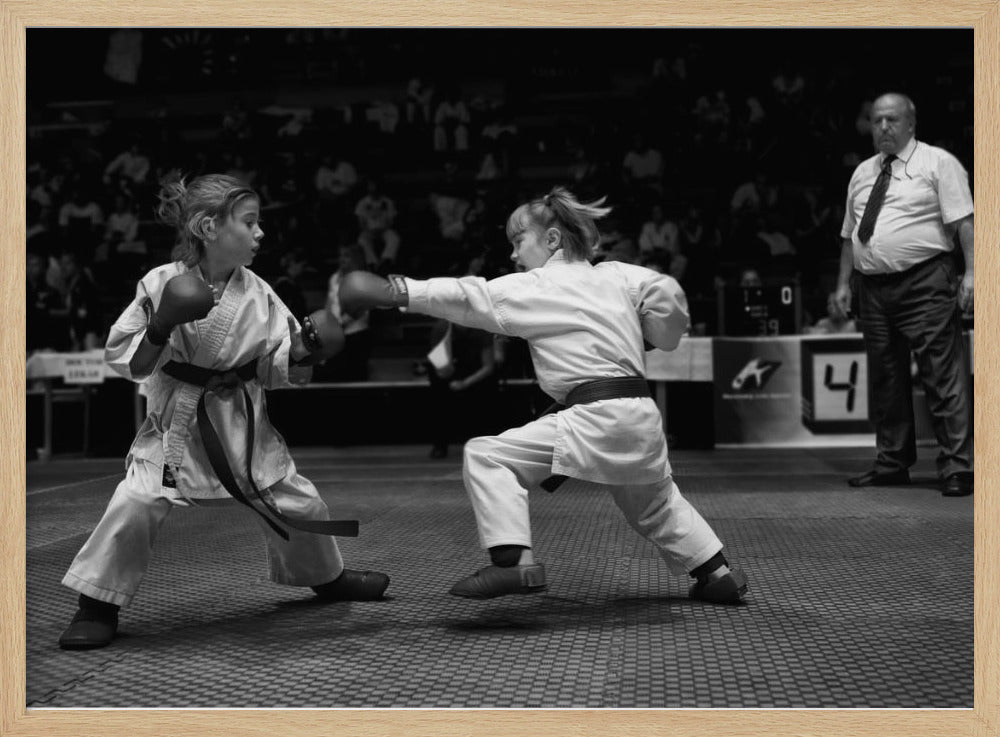 A black and white photograph of two young girls in a karate competition. They are wearing white gis, black belts, and boxing gloves. One girl is in the middle of throwing a punch at her opponent. In the background, a referee stands watching the match, and spectators are blurred in the stands. The image is presented within a silver-colored frame. Print