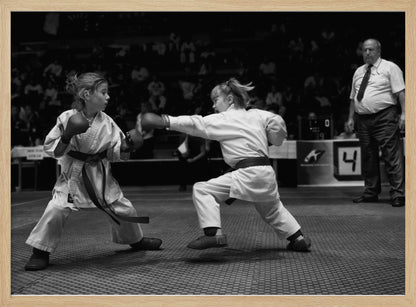 A black and white photograph of two young girls in a karate competition. They are wearing white gis, black belts, and boxing gloves. One girl is in the middle of throwing a punch at her opponent. In the background, a referee stands watching the match, and spectators are blurred in the stands. The image is presented within a silver-colored frame. Print