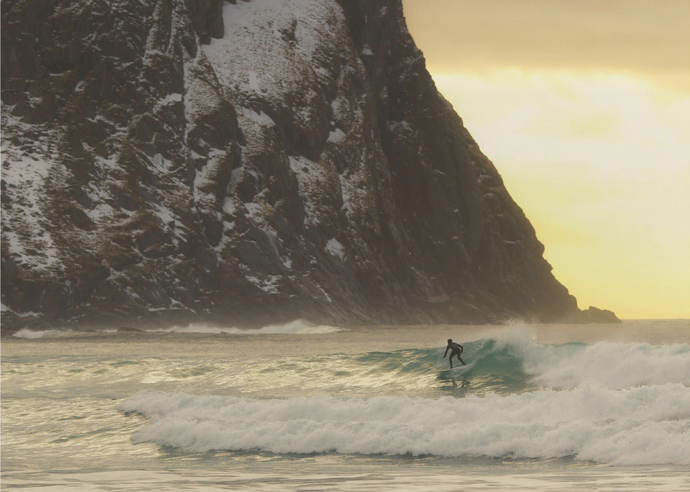 A framed photograph captures a lone surfer in a black wetsuit riding a turquoise wave. In the background, a massive, steep, snow-dusted mountain looms over the cold sea under a pale yellow, overcast sky. Print