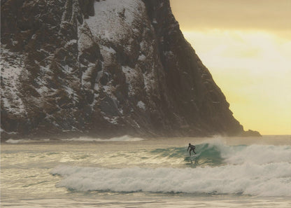 A framed photograph captures a lone surfer in a black wetsuit riding a turquoise wave. In the background, a massive, steep, snow-dusted mountain looms over the cold sea under a pale yellow, overcast sky. Print