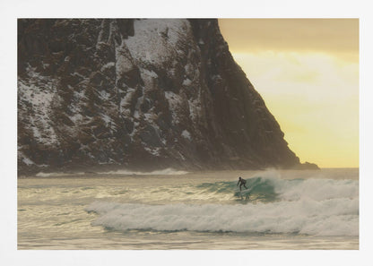 A framed photograph captures a lone surfer in a black wetsuit riding a turquoise wave. In the background, a massive, steep, snow-dusted mountain looms over the cold sea under a pale yellow, overcast sky. Print