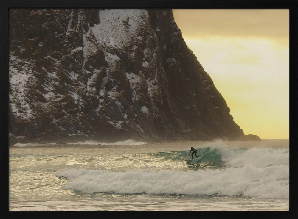 A framed photograph captures a lone surfer in a black wetsuit riding a turquoise wave. In the background, a massive, steep, snow-dusted mountain looms over the cold sea under a pale yellow, overcast sky. Print