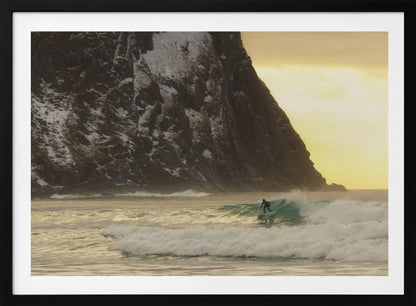 A framed photograph captures a lone surfer in a black wetsuit riding a turquoise wave. In the background, a massive, steep, snow-dusted mountain looms over the cold sea under a pale yellow, overcast sky. Print