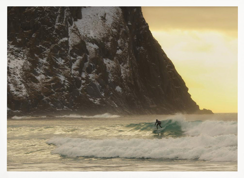 A framed photograph captures a lone surfer in a black wetsuit riding a turquoise wave. In the background, a massive, steep, snow-dusted mountain looms over the cold sea under a pale yellow, overcast sky. Print