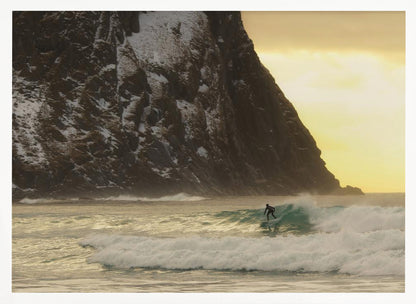 A framed photograph captures a lone surfer in a black wetsuit riding a turquoise wave. In the background, a massive, steep, snow-dusted mountain looms over the cold sea under a pale yellow, overcast sky. Print