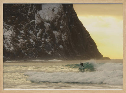 A framed photograph captures a lone surfer in a black wetsuit riding a turquoise wave. In the background, a massive, steep, snow-dusted mountain looms over the cold sea under a pale yellow, overcast sky. Print