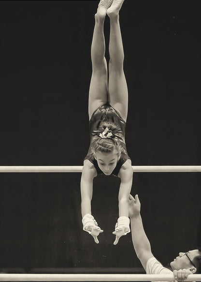 A dramatic black and white photograph of a young female gymnast performing a flawless handstand on the top of two parallel bars. Her body is perfectly vertical against a stark black background. Below, a male coach looks up at her with focused attention, his hand raised nearby, ready to spot her. Decor