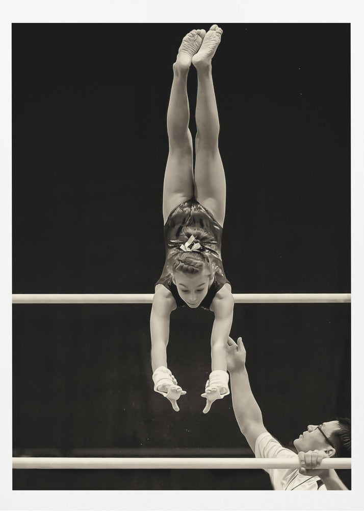 A dramatic black and white photograph of a young female gymnast performing a flawless handstand on the top of two parallel bars. Her body is perfectly vertical against a stark black background. Below, a male coach looks up at her with focused attention, his hand raised nearby, ready to spot her. Decor