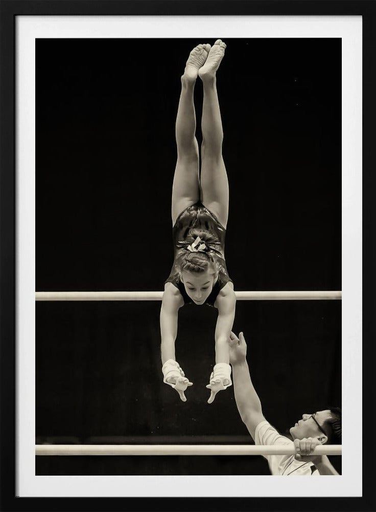 A dramatic black and white photograph of a young female gymnast performing a flawless handstand on the top of two parallel bars. Her body is perfectly vertical against a stark black background. Below, a male coach looks up at her with focused attention, his hand raised nearby, ready to spot her. Decor