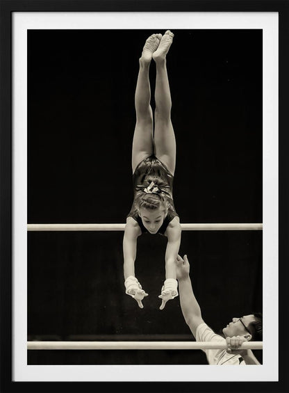 A dramatic black and white photograph of a young female gymnast performing a flawless handstand on the top of two parallel bars. Her body is perfectly vertical against a stark black background. Below, a male coach looks up at her with focused attention, his hand raised nearby, ready to spot her. Decor