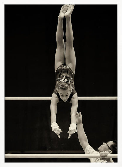 A dramatic black and white photograph of a young female gymnast performing a flawless handstand on the top of two parallel bars. Her body is perfectly vertical against a stark black background. Below, a male coach looks up at her with focused attention, his hand raised nearby, ready to spot her. Decor