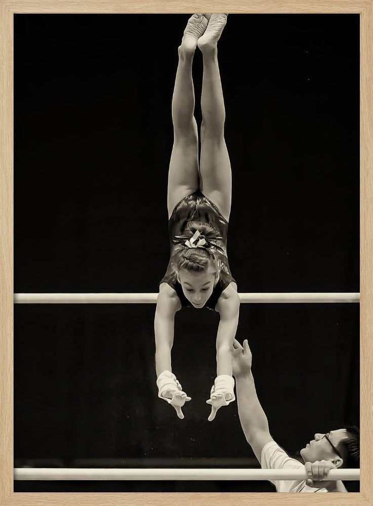 A dramatic black and white photograph of a young female gymnast performing a flawless handstand on the top of two parallel bars. Her body is perfectly vertical against a stark black background. Below, a male coach looks up at her with focused attention, his hand raised nearby, ready to spot her. Decor