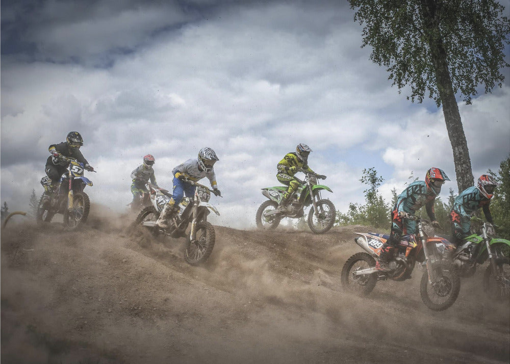 A dynamic action shot of several motocross riders racing up a dirt hill, kicking up a large cloud of dust. The riders are in full gear on colorful bikes, with a dramatic cloudy sky and a tall tree in the background. Print