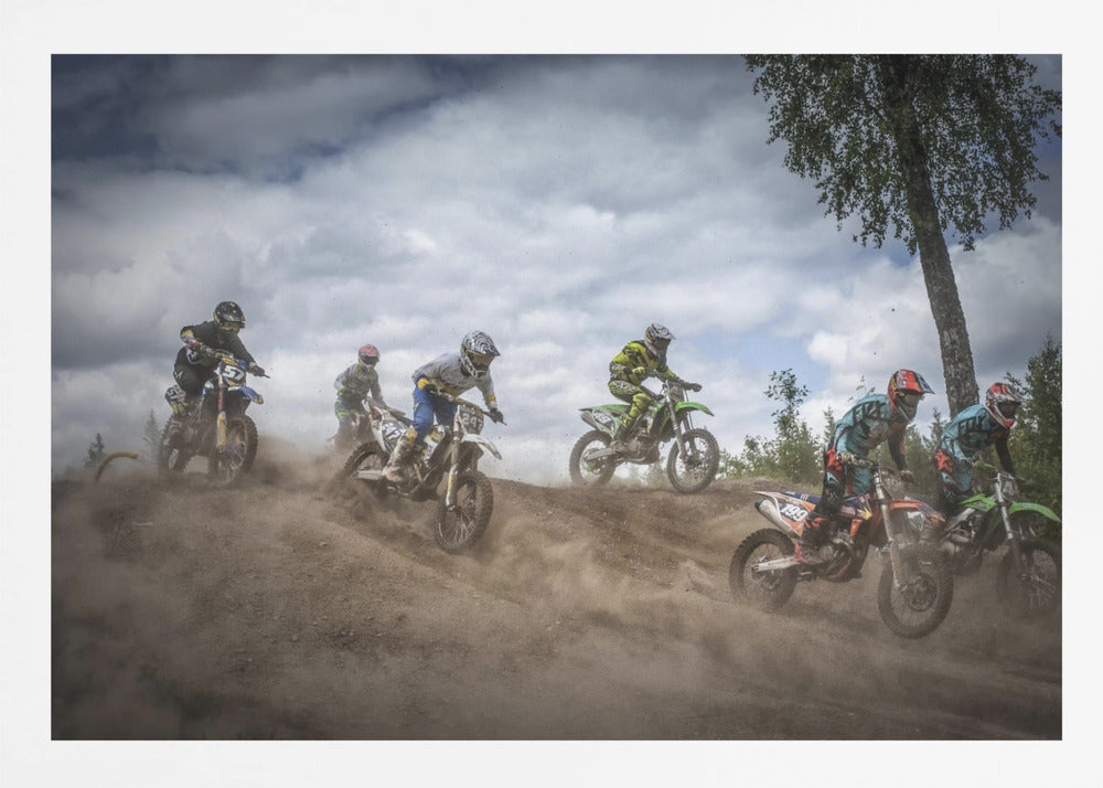 A dynamic action shot of several motocross riders racing up a dirt hill, kicking up a large cloud of dust. The riders are in full gear on colorful bikes, with a dramatic cloudy sky and a tall tree in the background. Print