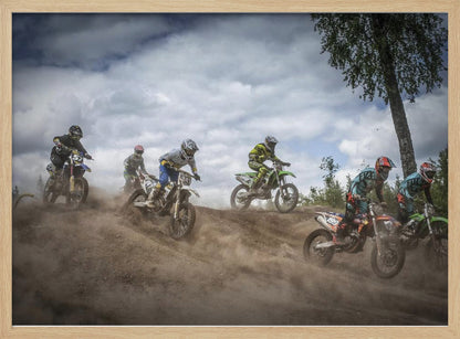 A dynamic action shot of several motocross riders racing up a dirt hill, kicking up a large cloud of dust. The riders are in full gear on colorful bikes, with a dramatic cloudy sky and a tall tree in the background. Print
