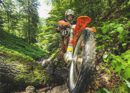 A motocross rider on an orange dirt bike tackles a muddy, wooded trail, maneuvering around a moss-covered log in a lush green forest, seen from a low-angle perspective. Decor