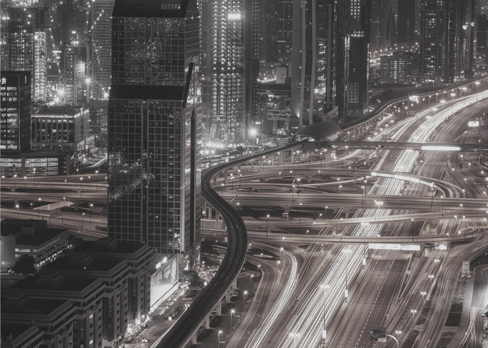 A dynamic black and white long-exposure photograph of a sprawling metropolis at night, viewed from above. Gleaming skyscrapers fill the background, while a complex interchange of highways with streaks of light from traffic dominates the foreground, with an elevated train line weaving through the city. Wall Art